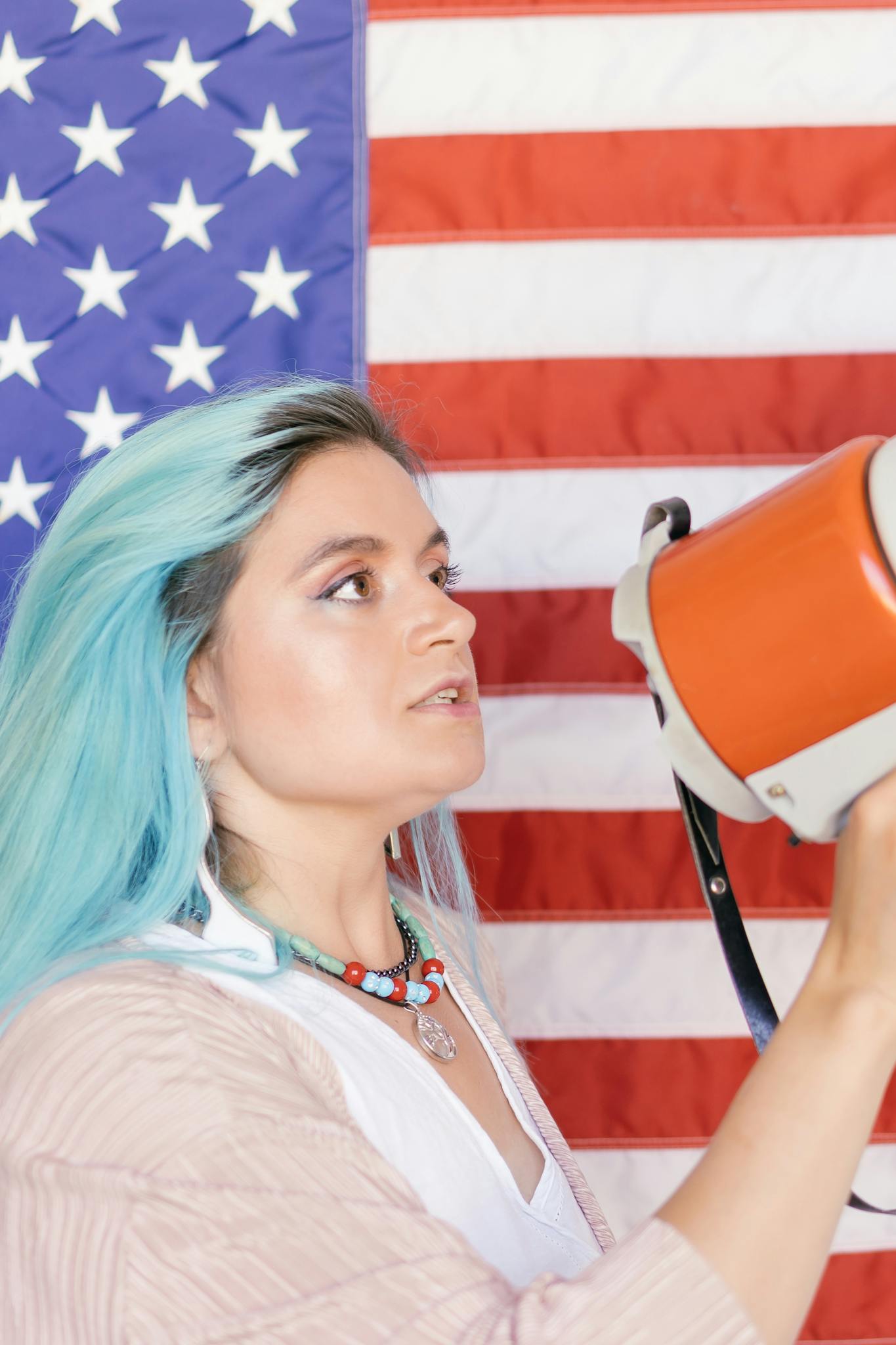 Caucasian woman with blue hair holding megaphone in front of American flag.