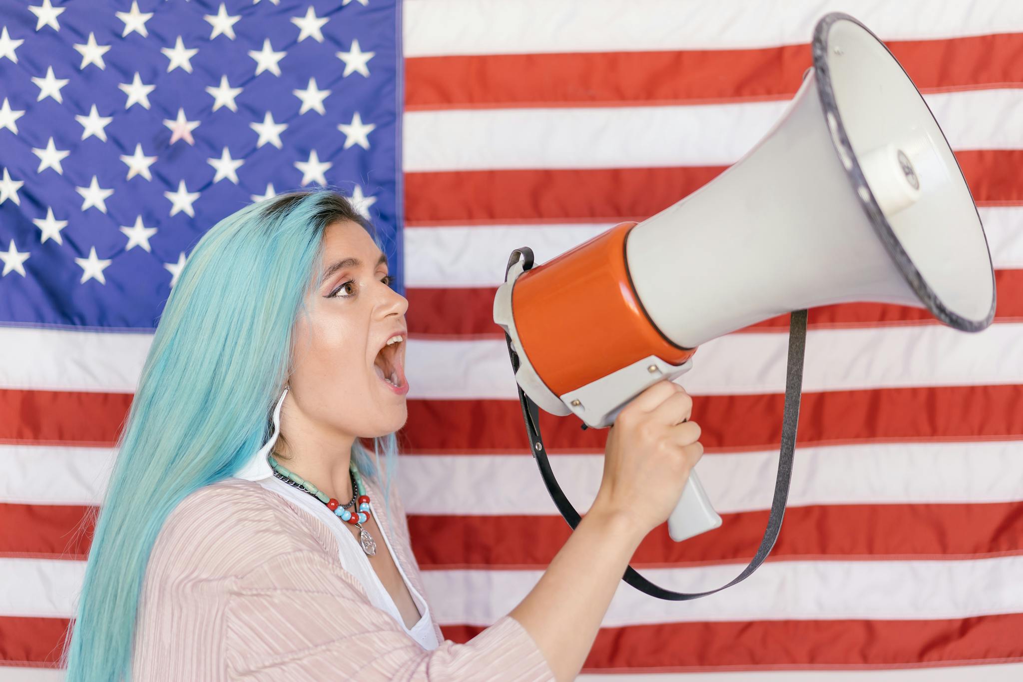 Woman with blue hair shouting through a megaphone in front of an American flag.